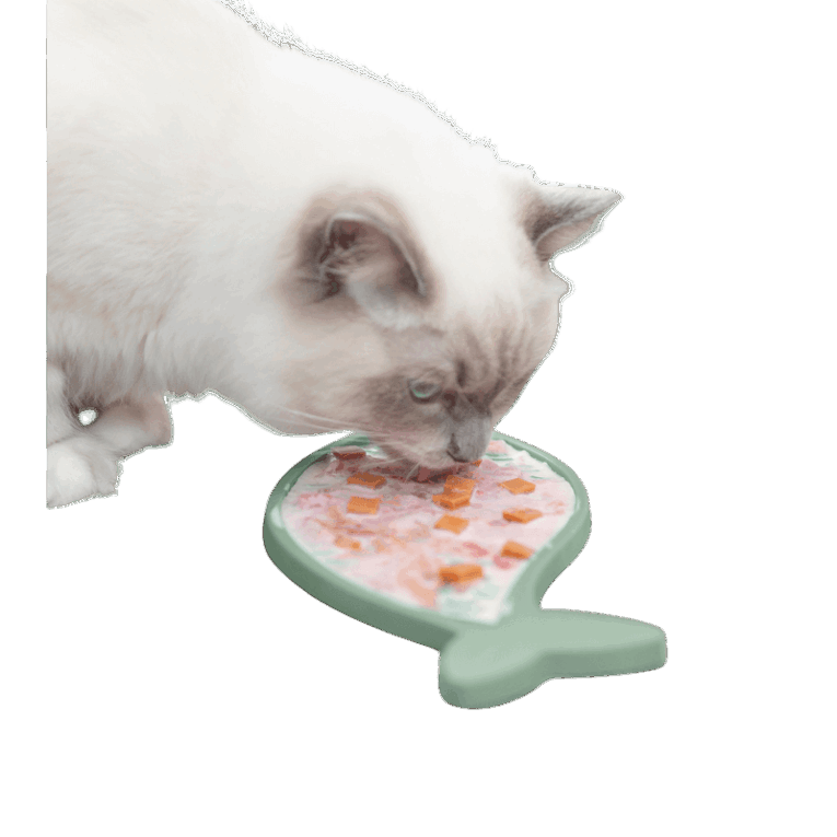Un gato de raza Ragdoll (o similar) lamiendo el mantel Trixie Lick'n'Snack de silicona con forma de pez. El tapete está cargado con una mezcla de comida húmeda y trozos de vegetales.