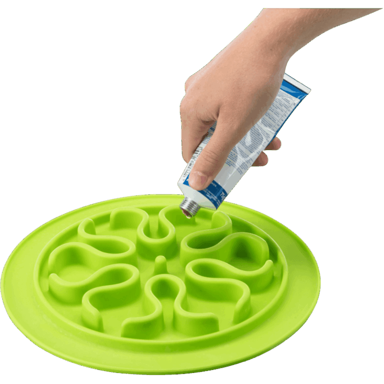 Primer plano de una mano de persona rellenando el laberinto del comedero slow feed redondo de 24cm con un snack cremoso o paté directamente de un tubo. Se aprecia el diseño de ondas en el comedero verde.