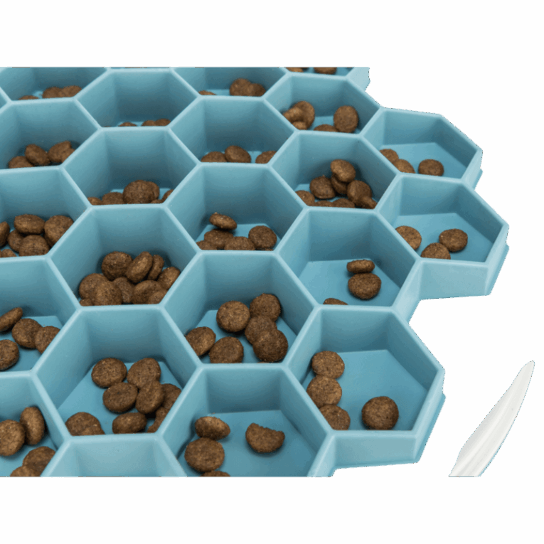 Vista de cerca del comedero Trixie Colmena azul de 30cm, con croquetas de pienso dispersas individualmente en las celdas hexagonales. Muestra cómo las celdas separan el alimento.