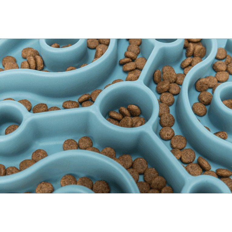 Primer plano con zoom del patrón de laberinto curvo del mantel Slow Feeding Trixie azul. Se ven claramente las croquetas de pienso agrupadas en los pozos y dispersas en los canales del centro.