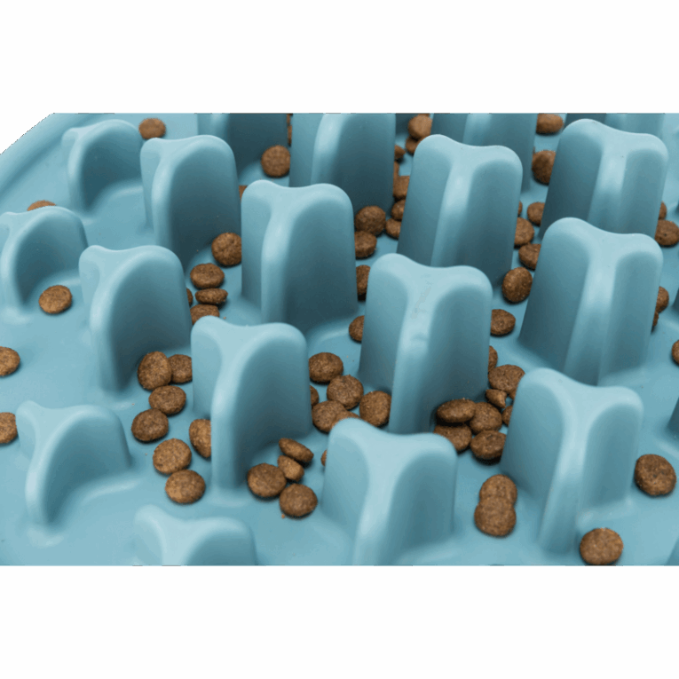 Primer plano de la alfombrilla Trixie Slow Feeding de 35cm, color azul. Muestra las croquetas de pienso dispersas entre los pilares de silicona, enfatizando el tamaño y la dificultad de la ingesta.